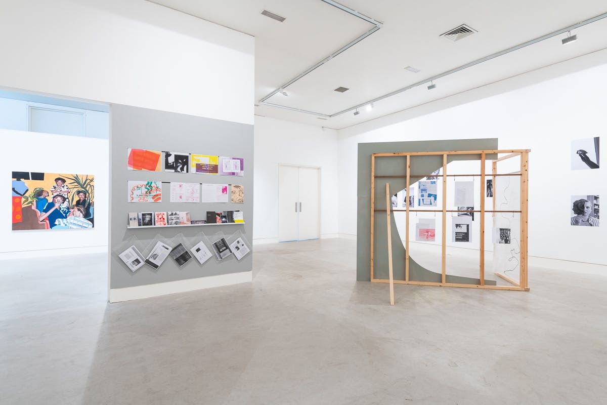 From the left to the right. Their is a painting of a happy family. In the middle of the image is a grey wall with around 40 publications pinned to it. On the right hand side is a timber construction with more publications hanging from it. The gallery is light and bright. 