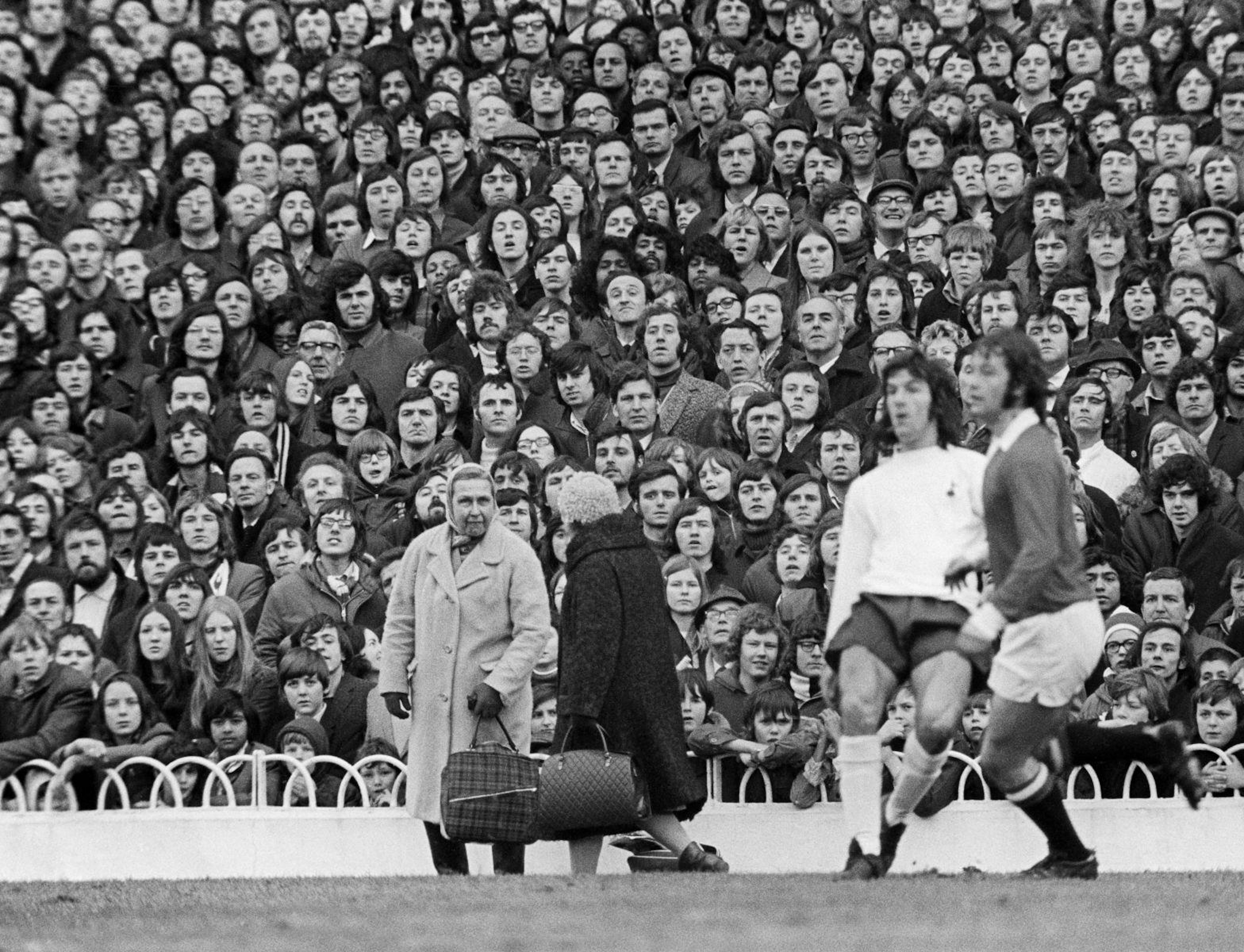 It is a black and white photograph of two footballers engaged in action. The camera points upwards and focuses on the reaction of the crowd who remain rapt in attention. In the middle ground two older ladies are chatting, obvious to the action. 