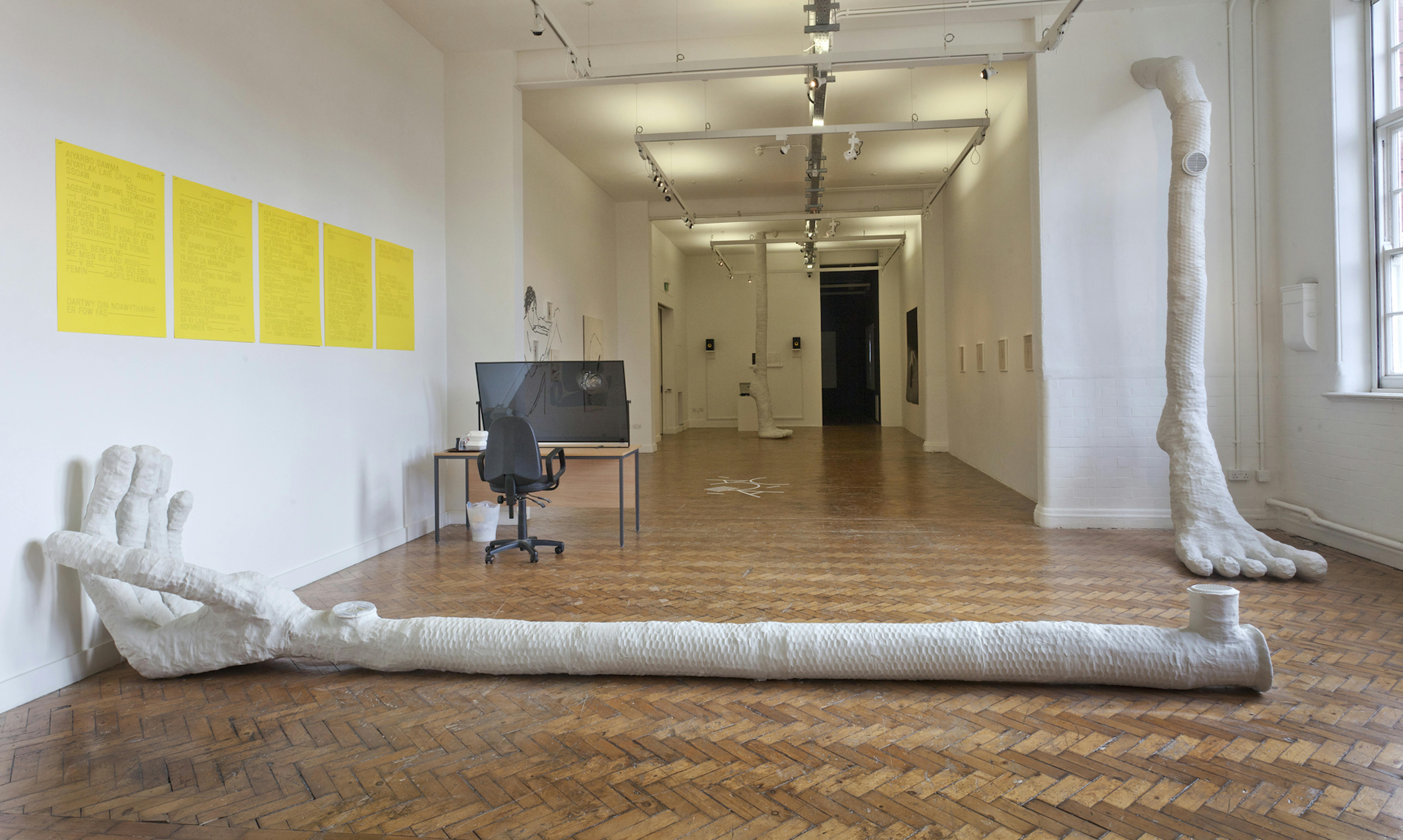 A large white hand made from plaster lies on the floor and a large white foot made in plaster appears from the wall. They frame a series of yellow posters with lettering on it. The yellow posters form a type of enigmatic poetry with fragmented words. 