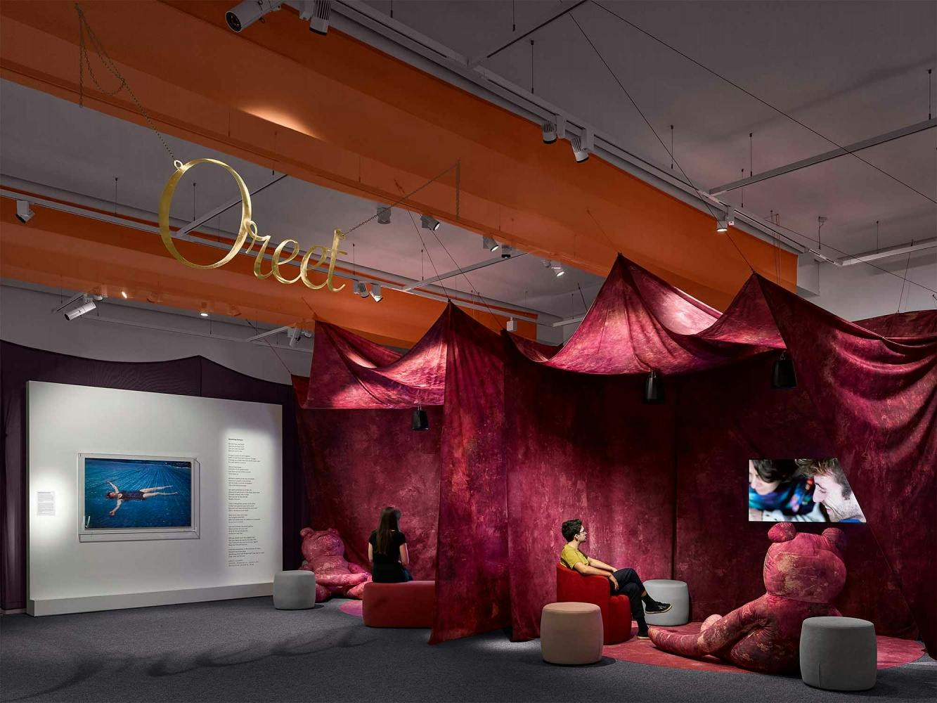 The installation includes photographs on walls of Jo Spence and lots of draped fabric creating a comforting environment. On the ceiling, Oreet Ashery's name is spelled out in gold. In the background is a large photograph of Jo Spence floating in water. In the foreground people watch TV screens surrounded by giant teddy bears. 