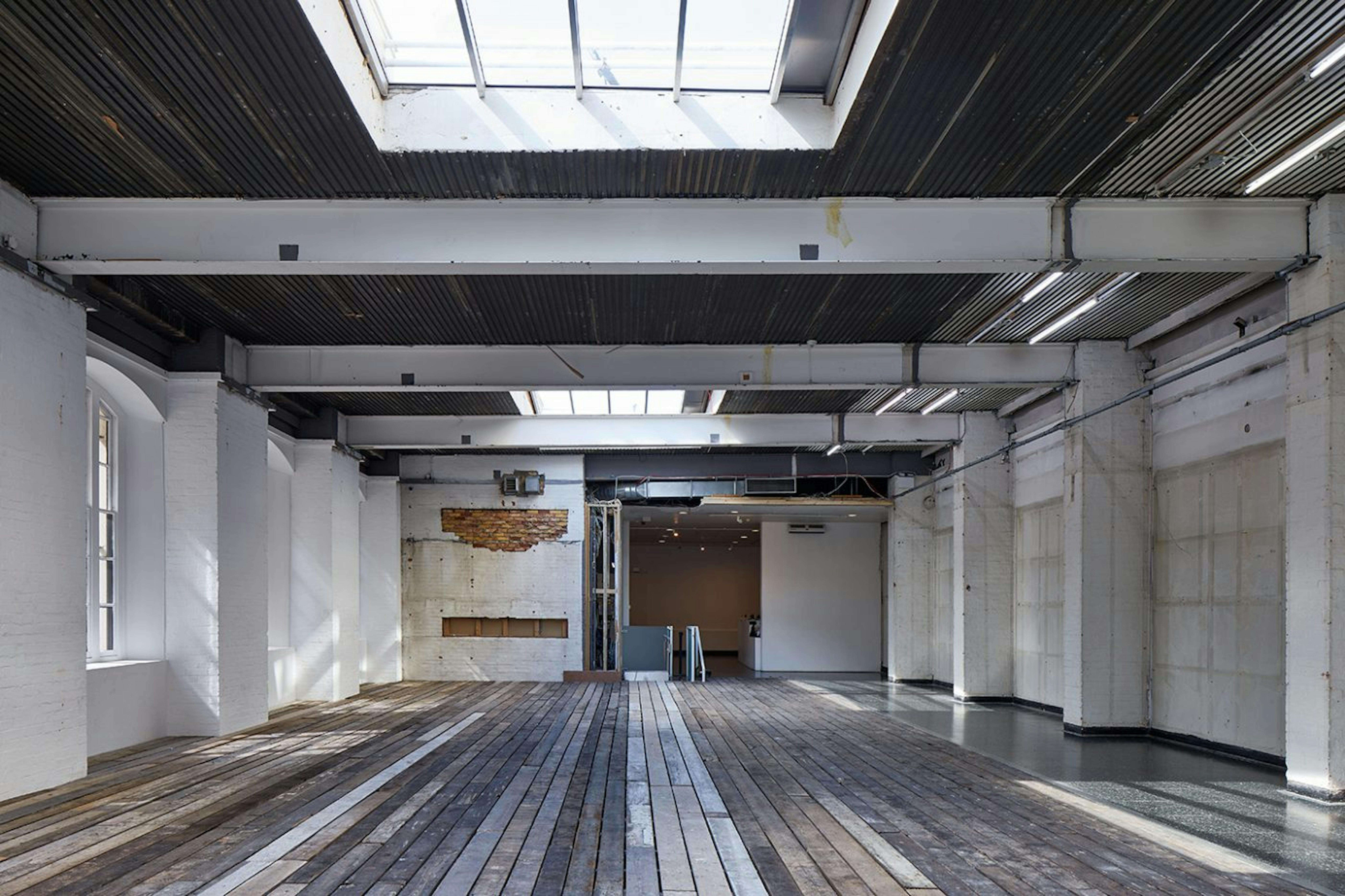 An empty gallery with everything stripped back. The floor is made from old wooden beams and the walls are stained and unflustered. Natural light seeps into the space through windows on the left hand side and top skylight. 