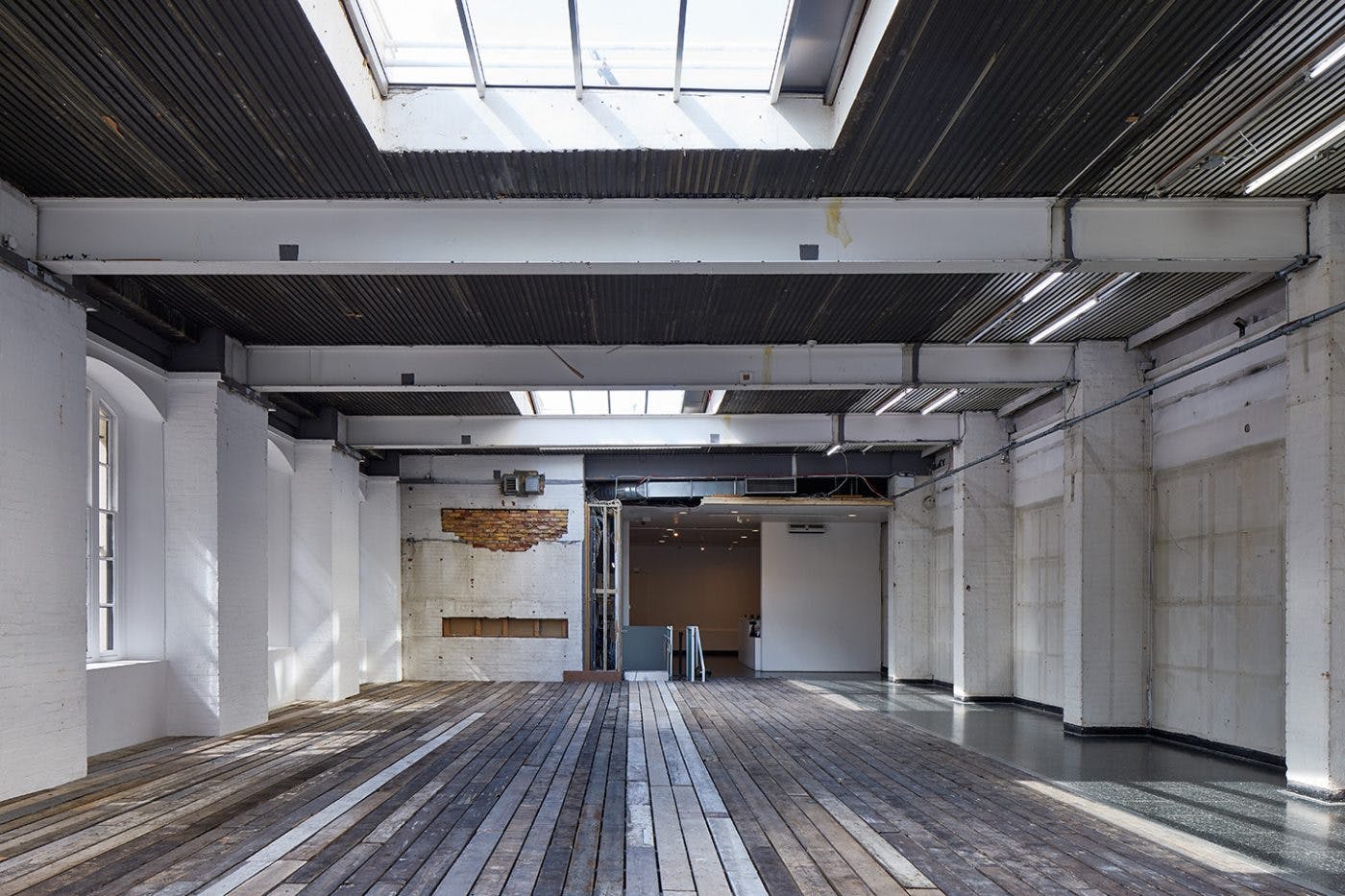 An empty gallery with everything stripped back. The floor is made from old wooden beams and the walls are stained and unflustered. Natural light seeps into the space through windows on the left hand side and top skylight.