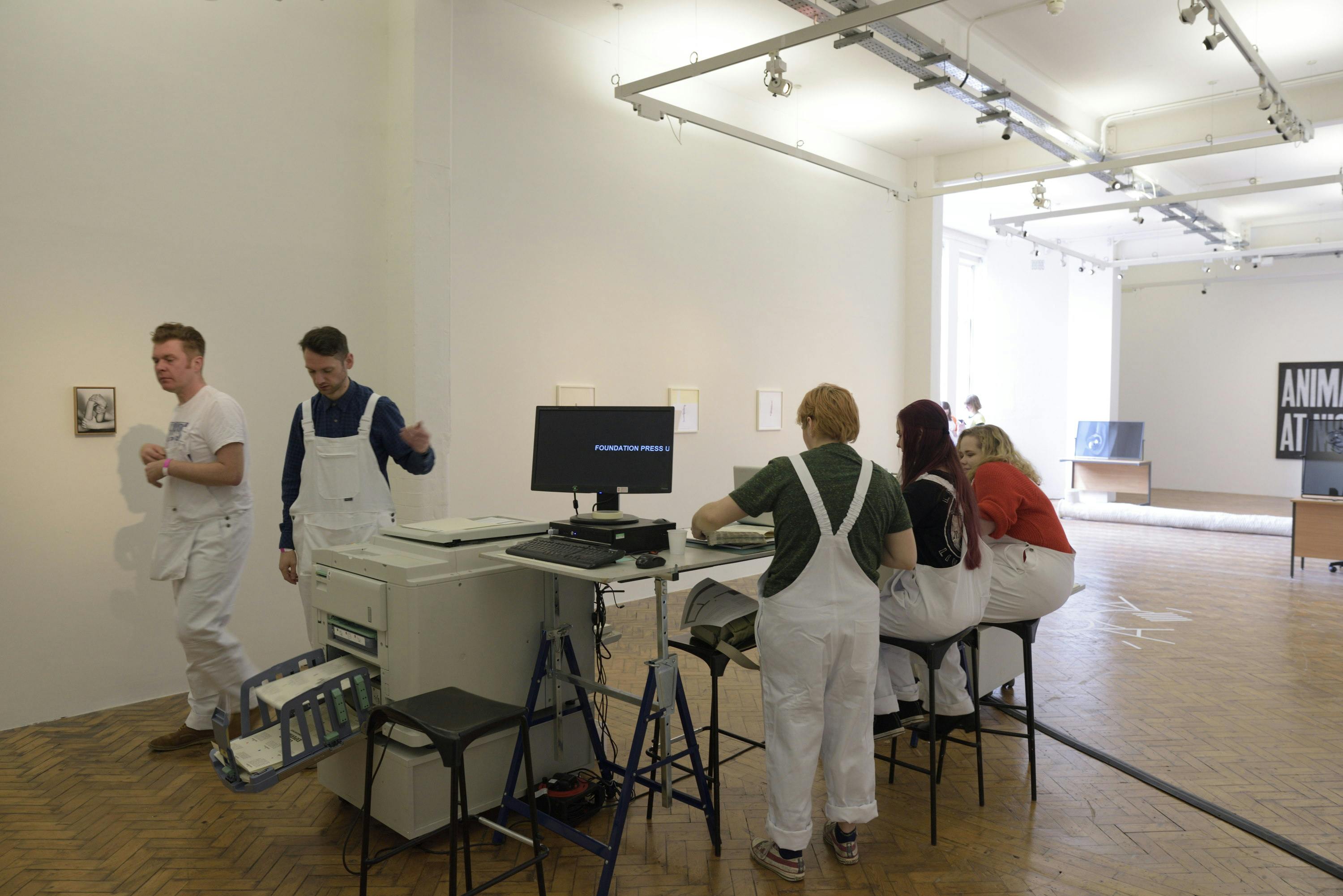 A workshop is taking place in the gallery. A group of students dressed in white uniforms surround a printer. 