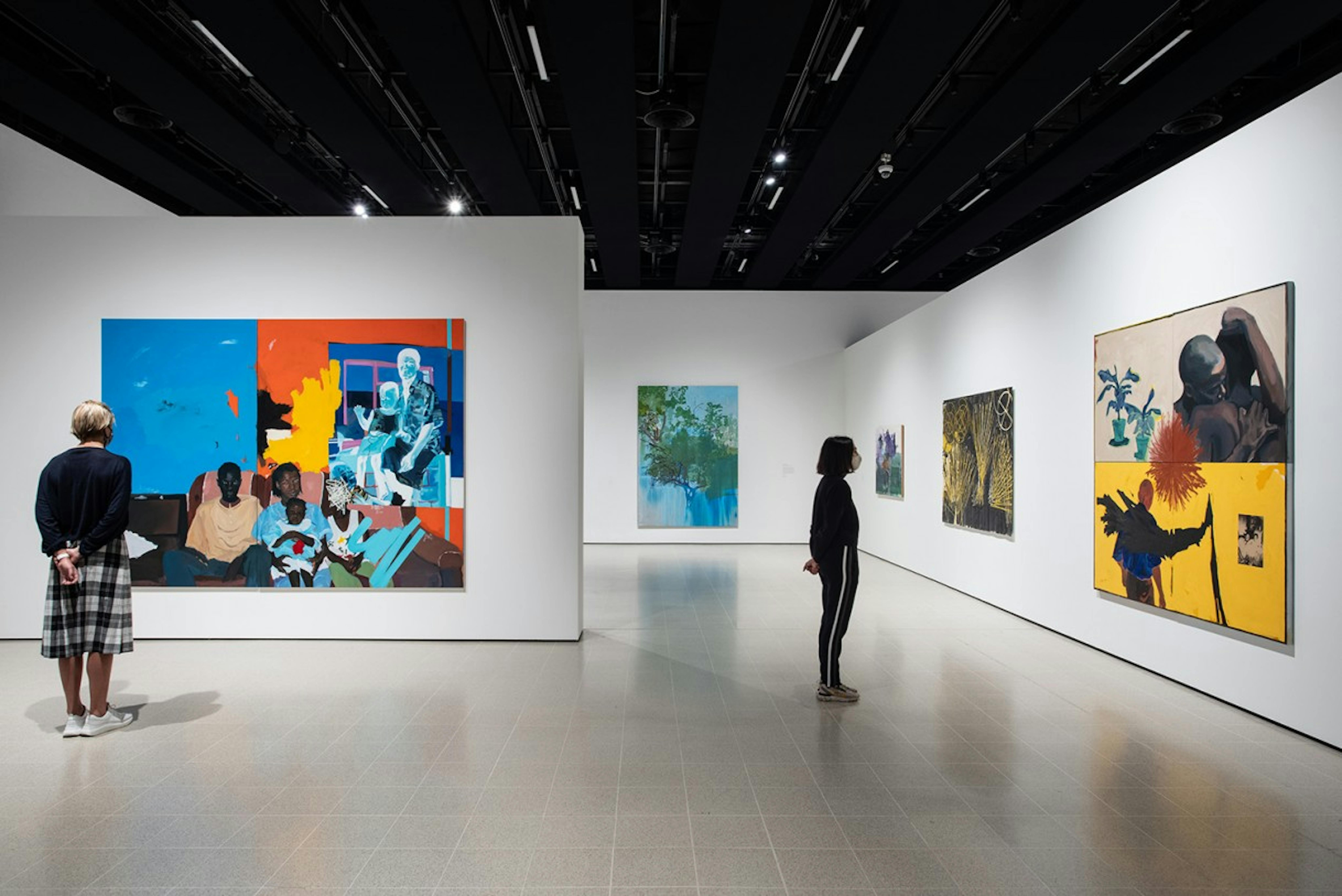 An installation of a gallery with large figurative and colourful paintings throughout. Two figures stand and look at the paintings providing a sense of scale. 