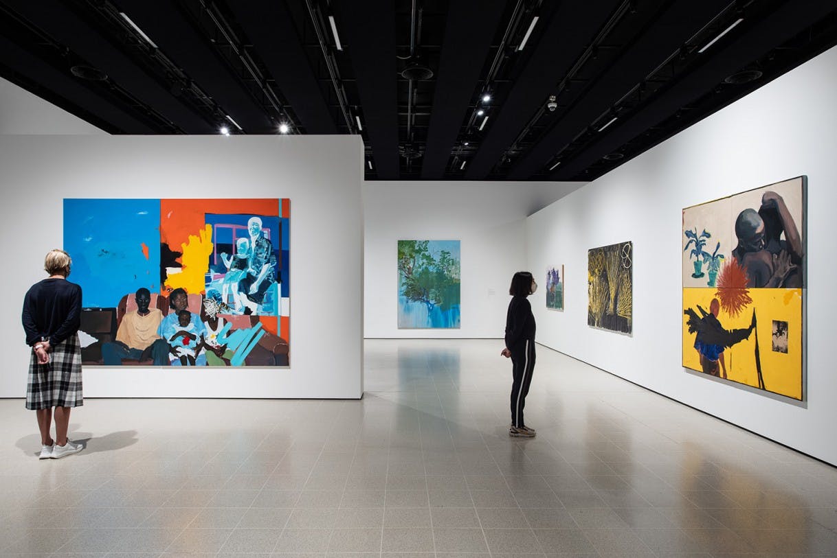 An installation of a gallery with large figurative and colourful paintings throughout. Two figures stand and look at the paintings providing a sense of scale. 