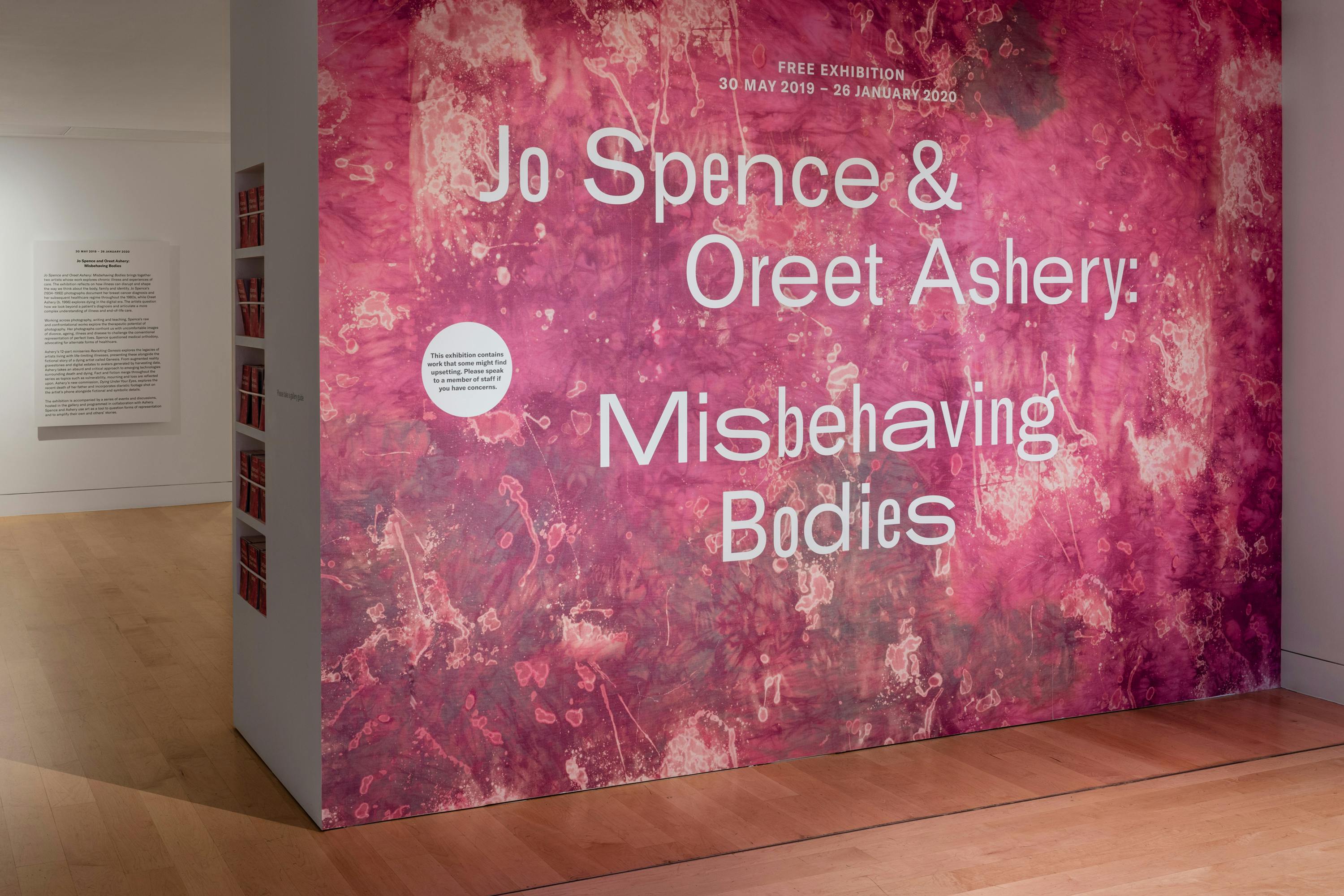 The wall has the title of the exhibition Jo Spence and Oreet Ashery Misbehaving Bodies and is wallpapered with deep red tie dye. 