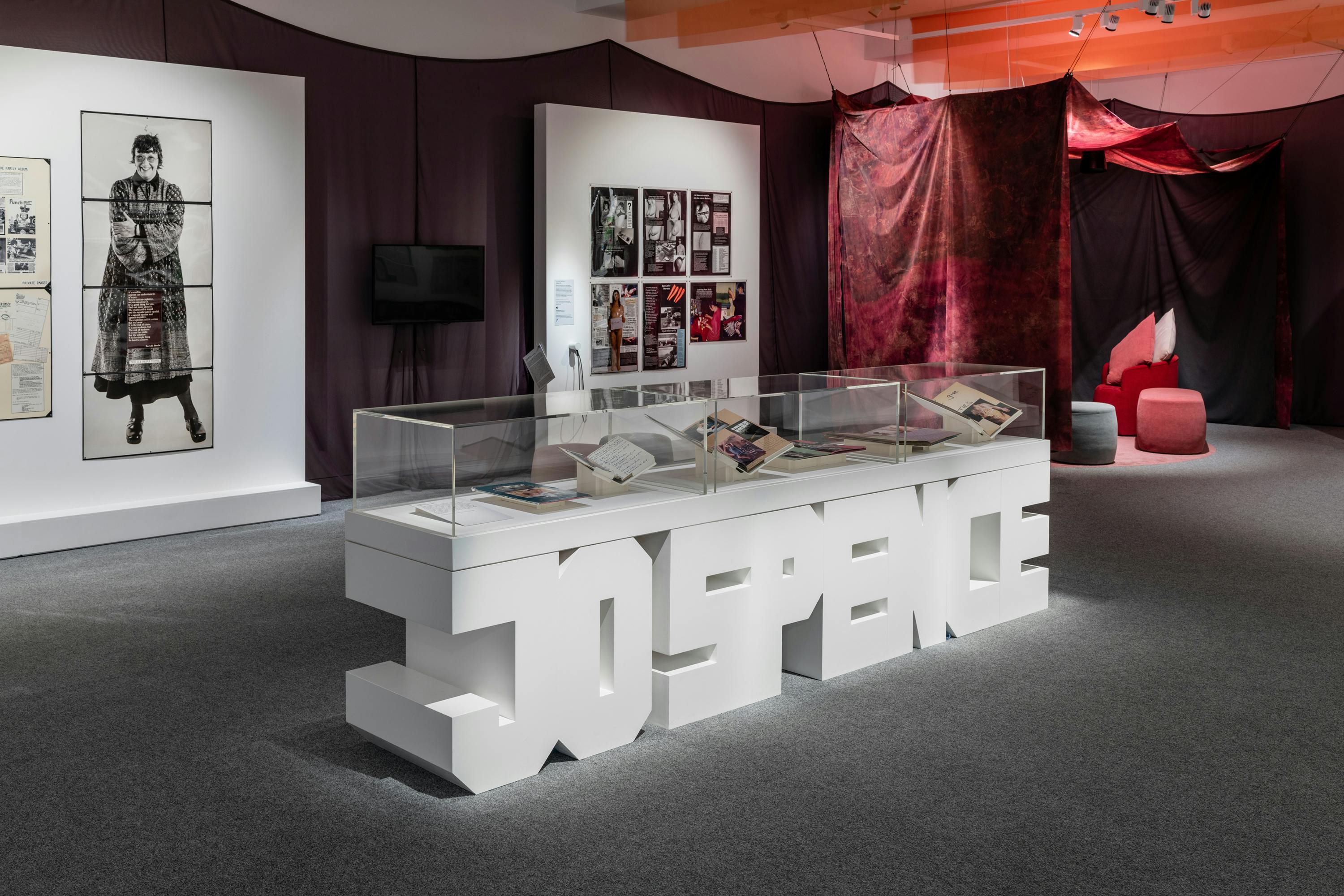 The installation includes large photographs and montages of Jo Spence on white walls. In the foreground of the image a vitrine is shaped in Jo Spence's name and contains notebooks and diaries. The gallery walls and ceiling are covered in peach coloured and dark brown fabrics creating a comforting environment.