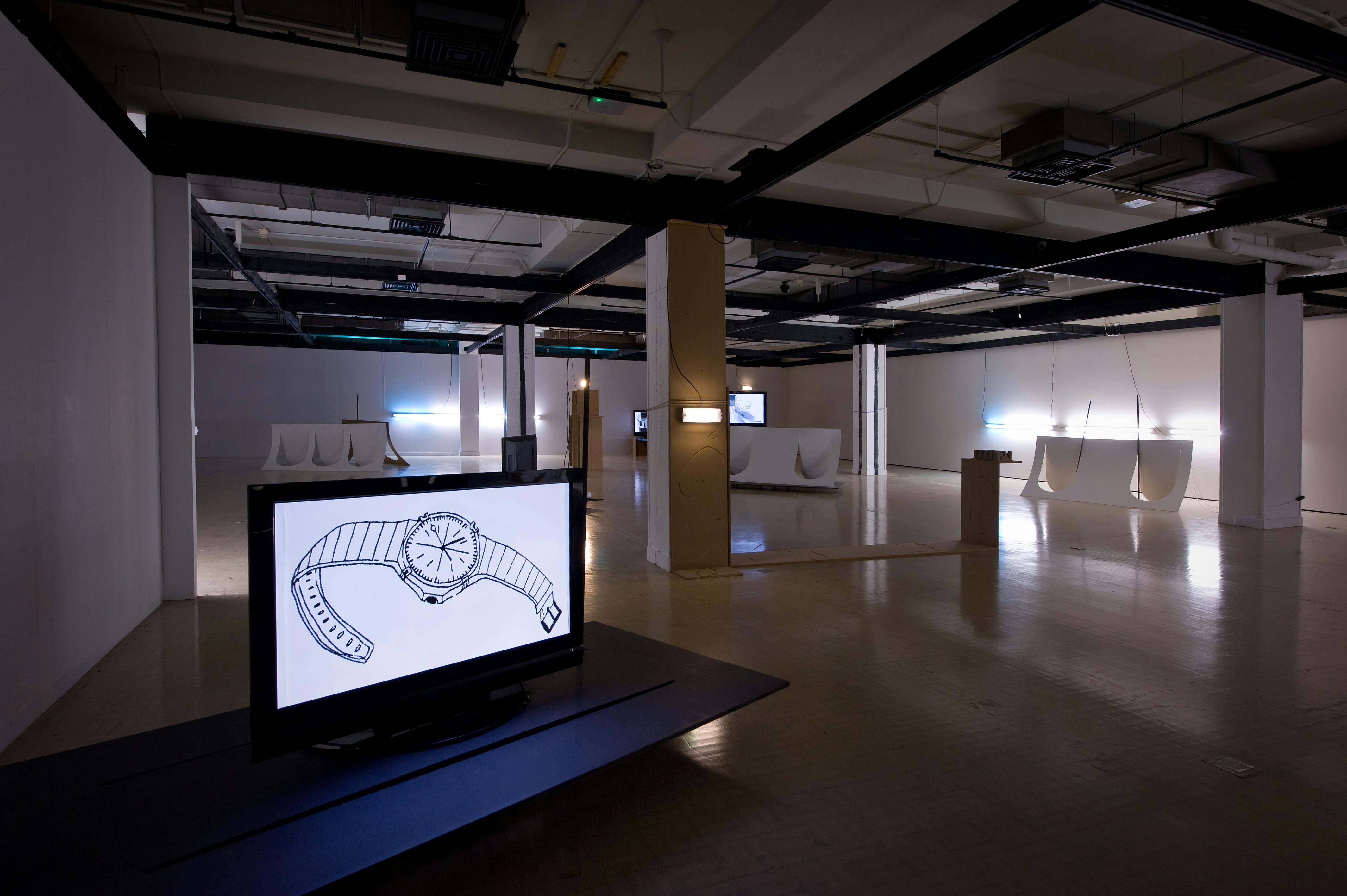 An installation in a darkened gallery with flat screen TV monitors on mobile platforms. The screen in the foreground has an image of a watch.
