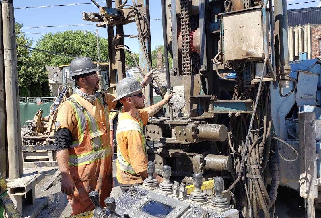 Two workers on site in front of a large drill. One worker is teaching the other.