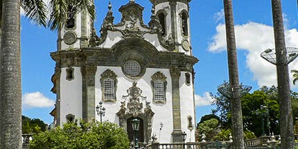 Imagem da Igreja de São Francisco de Assis em São João del Rei, Minas Gerais. A foto mostra a fachada branca da igreja, rodeada de árvores e palmeiras em um dia ensolarado.