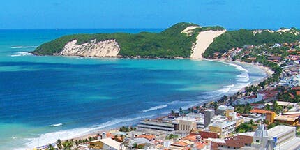 Imagem da praia de Natal, no Rio Grande do Norte, em dia ensolarado. A foto mostra a costa com o mar azul, vegetação e casa ao redor.