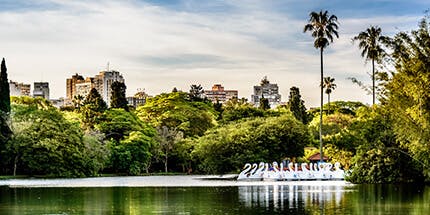 Imagem de parque em Porto Alegre, no Rio Grande do Sul, em dia ensolarado. A foto mostra o lago, com árvores ao redor e paisagem de prédios da cidade ao fundo.
