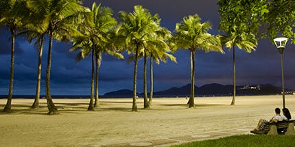 Imagem da Praia de São Vicente, em São Paulo, ao entardecer. A foto mostra o céu azulado, com palmeiras na areia da praia e montanhas ao fundo.