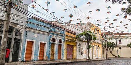 Imagem da Rua do Bom Jesus, no centro antigo do Recife. A foto mostra casas coloridas em dia ensolarado, com guarda-chuvas tradicionais da região pendurados sobre a rua