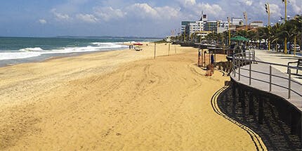 Imagem da praia de Macaé, no Rio de Janeiro, em dia ensolarado. A foto mostra a orla, com a areia e o mar e paisagem de prédios ao fundo.