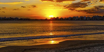Imagem da praia de Guarapari, no Espírito Santo, ao pôr do sol. A foto mostra o céu alaranjado refletindo no mar, com prédios ao fundo.