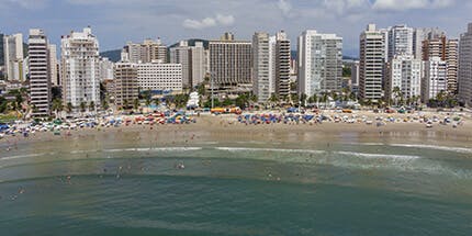 Imagem panorâmica da cidade do Guarujá, em São Paulo. A foto mostra a praia em dia ensolarado, com visão em primeiro plano do mar e da areia com turistas e prédios ao fundo.