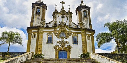 Imagem da Igreja de São Francisco de Assis, em Ouro Preto, São Paulo. A foto mostra a vista da igreja em dia ensolarado, que é cercada por montanhas e vegetação.