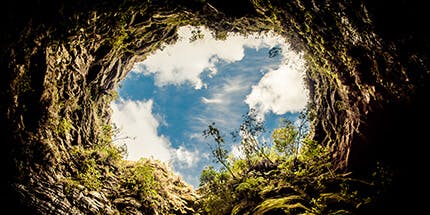 Imagem do Buraco do Padre em Ponta Grossa, no Paraná. A foto mostra a visão de dentro do buraco, com vista do céu azul com nuvens, e rochas e vegetação ao seu redor.