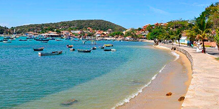Imagem da orla da praia de Armação de Búzios, no Rio de Janeiro. A foto mostra um dia ensolarado, o mar com água cristalina com pequenos barcos de turismo e a paisagem da cidade e montanhas ao fundo.