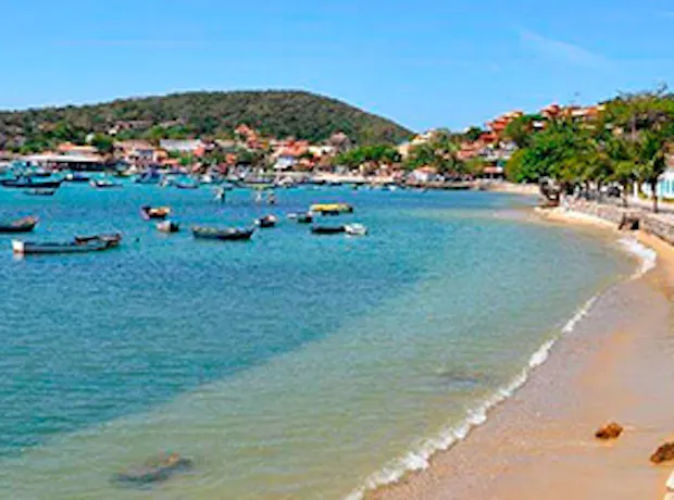 Imagem da orla da praia de Armação de Búzios, no Rio de Janeiro. A foto mostra um dia ensolarado, o mar com água cristalina com pequenos barcos de turismo e a paisagem da cidade e montanhas ao fundo.