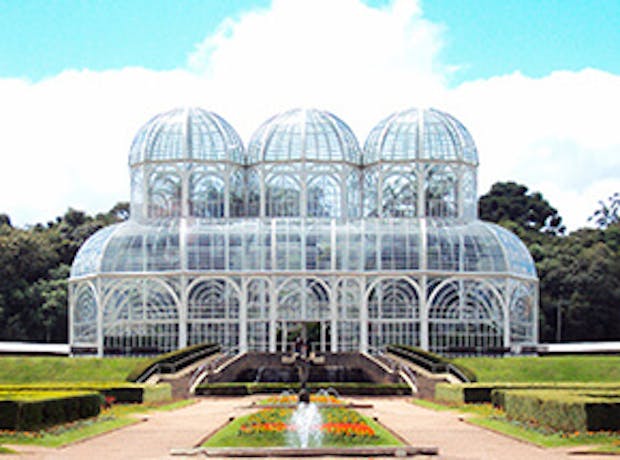 Imagem do Jardim Botânico de Curitiba, no Paraná, em dia ensolarado. A foto mostra a estrutura de vidro da estufa, cercada por arbustos, flores e árvores.