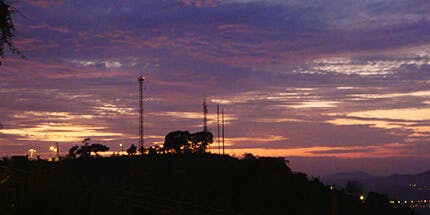 Imagem da cidade de Volta Redonda, no Rio de Janeiro, ao entardecer. A foto mostra a silhueta de prédios e árvores da cidade contra o céu alaranjado.