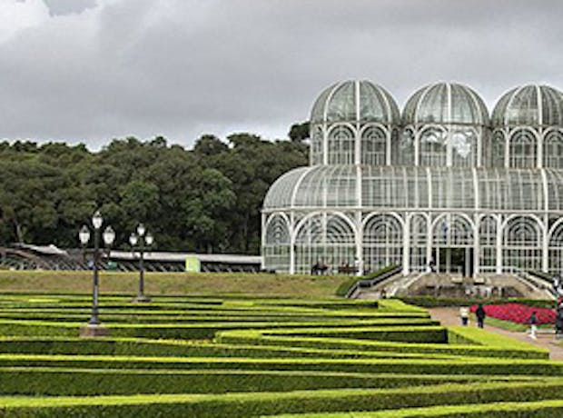 Imagem do Jardim Botânico de Curitiba, no Paraná, em dia ensolarado. A foto mostra a estrutura de vidro da estufa, cercada por arbustos, flores e árvores.
