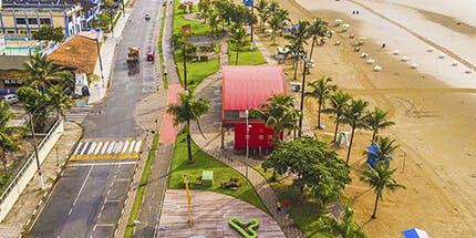 Imagem da praia de Bertioga, em São Paulo, em dia ensolarado. A foto mostra a orla, com quiosques e palmeiras entre a cidade.