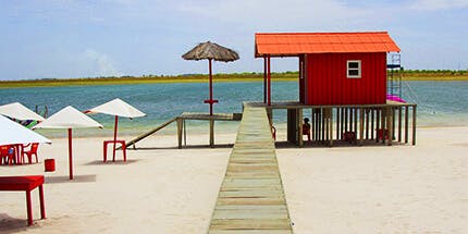 Imagem da praia em Boa Vista, em Roraima, em dia ensolarado. A imagem mostra um quiosque com mesas ao redor, com o mar ao fundo.