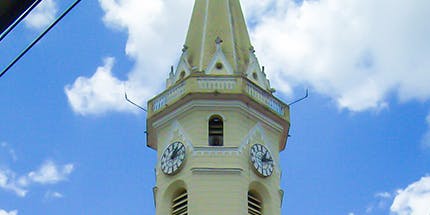 Imagem da Igreja de Sant'Ana em Lavras, Minas Gerais. A foto mostra uma de suas torres, que possui relógios ao seu redor, em dia ensolarado.