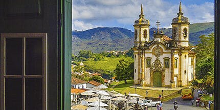 Imagem da Igreja de São Francisco de Assis, em Ouro Preto, São Paulo. A foto mostra a vista da igreja em dia ensolarado, que é cercada por montanhas e vegetação.