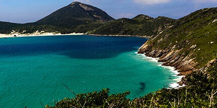 Imagem da costa de Arraial do Cabo, Rio de Janeiro, em dia ensolarado. A foto mostra o mar cristalino, com montanhas e vegetação natural local.