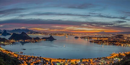 Imagem panorâmica da costa de Niterói, no Rio de Janeiro, ao entardecer. A imagem mostra o mar, com montanhas ao fundo e parte da cidade iluminada.