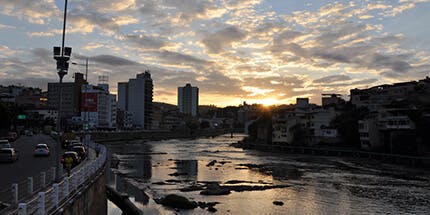 Imagem do Rio do Itapemirim, que cruza a cidade Cachoeiro do Itapemirim, no Espírito Santo. A foto ilustra o entardecer da cidade, com o reflexo do céu alaranjado no rio, e a paisagem de prédios ao fundo.