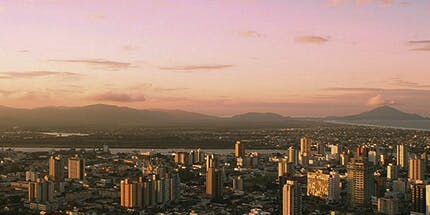 Imagem panorâmica da cidade de Itajaí, em Santa Catarina, ao amanhecer. A foto mostra o céu rosado da manhã com a paisagem de prédios da cidade com montanhas ao fundo.