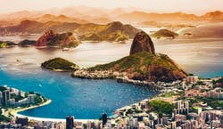 A imagem mostra uma visão panorâmica da cidade do Rio de Janeiro, com seu mar azul, montanhas em destaque e a orla da cidade ao entardecer, com tons alaranjados.