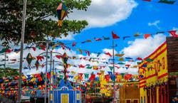 Imagem de celebração de São João. A foto mostra a praça de uma cidade, cercada de casas e estabelecimentos coloridos, com postes decorados com bandeirinhas e balões.
