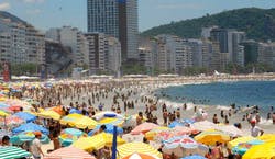 Imagem de praia em dia ensolarado. A foto mostra aorla da praia com inúmeros visitantes agrupados na areia com seus guarda-sóis, com prédios da cidade ao fundo.
