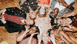 Roda de 8 pessoas deitadas no chão sorrindo enquanto confetes são jogados em celebração do carnaval.