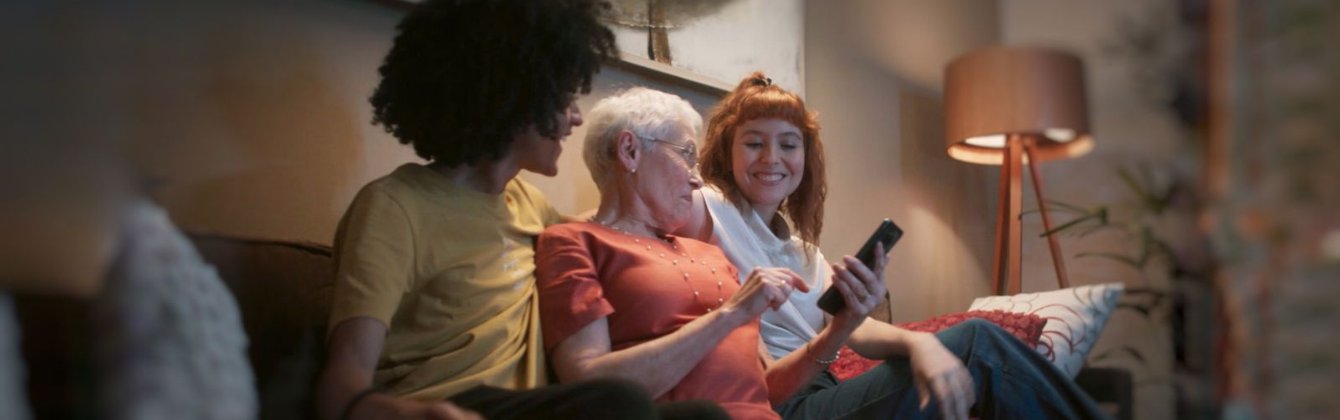 Imagem de capa da página. A foto mostra um homem negro jovem com uma camiseta amarela sentado ao lado de uma senhora de cabelos brancos, óculos e blusa vermelha e uma mulher jovem de cabelos ruivos e camiseta branca. Os três estão sentados em um sofá olhando para um celular preto enquanto acessam o Aplicativo da DeÔnibus.