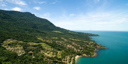 Imagem panorâmica da costa de Ilhabela em São Paulo. Em dia ensolarado, a água é cristalina e a paisagem mostra montanhas cobertas com a vegetação local.