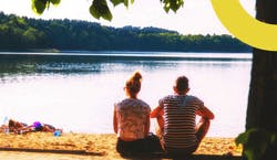 Homem e mulher sentados no chão à beira de um rio observando o pôr do sol.