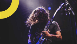 Imagem de homem com cabelo preto ondulado tocando uma guitarra preta durante show de rock.