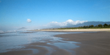 Imagem da praia de Boracéia, São Paulo, em dia ensolarado. A foto mostra a areia após a passagem de uma onda, o céu está azul e a paisagem tem montanhas e árvores ao fundo.