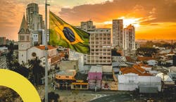 Imagem de bandeira do Brasil hasteada diante de uma cidade com prédios ao fundo durante o pôr do sol.