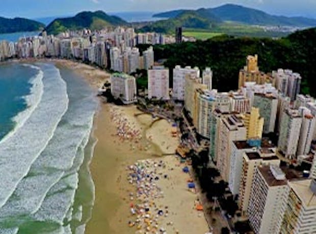 Imagem panorâmica da costa da cidade de Florianópolis, em Santa Catarina. A foto mostra um dia ensolarado com o mar azulado com ondas na praia, prédios e montanhas ao fundo.