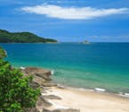 Imagem da praia de São Sebastião em dia ensolarado. A praia tem água cristalina com montanhas ao fundo e vegetação local ao redor de sua costa.