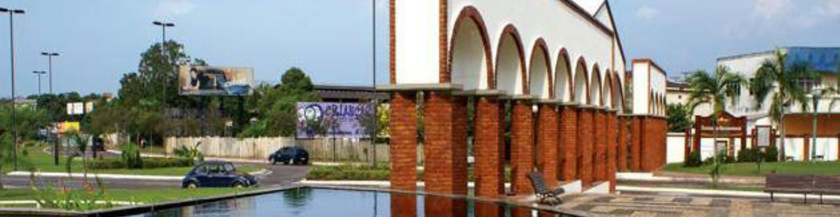 Pórtico do Parque da Matternidade. O pórtico é feito de tijolo aparente e na sua frente há uma lago. Ao redor, na rua pavimentada, tem dois carros estacionados.