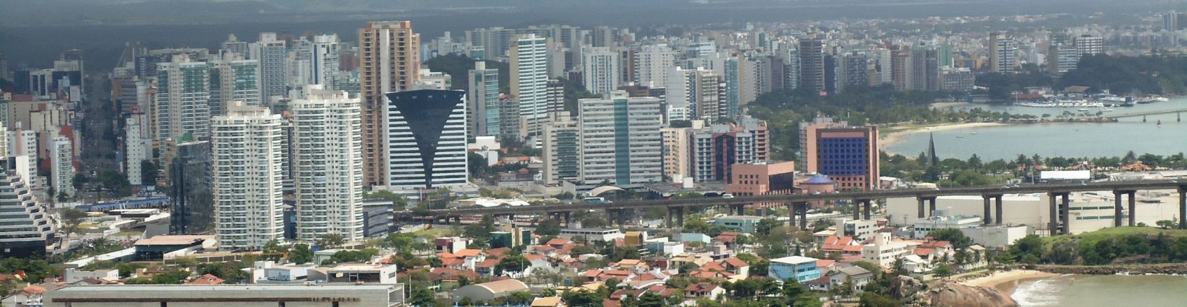 Vista panorâmica da área urbana da cidade de Vitória no Espírito Santo. Na imagem, casas e prédios ganham destaque, enquanto ao lado direito há uma parte da praia.